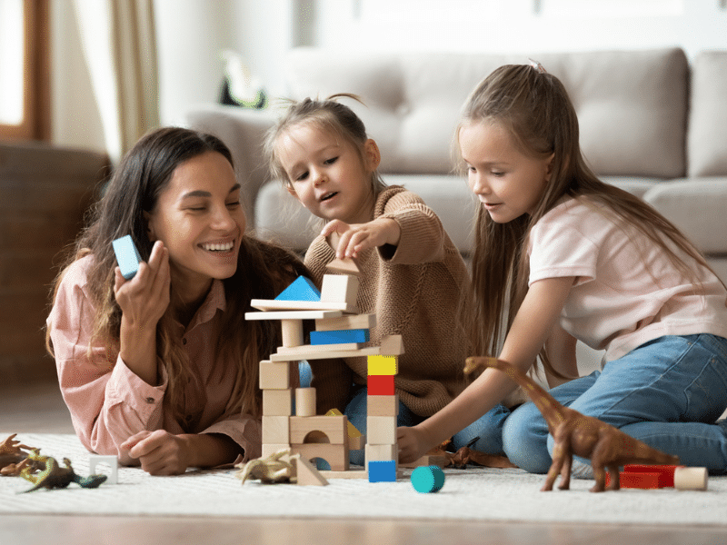 Happy young mum babysitter helping playing game with little kids daughters on floor, female nanny mother and cute small children sisters building castle of wooden blocks having fun with toys at home